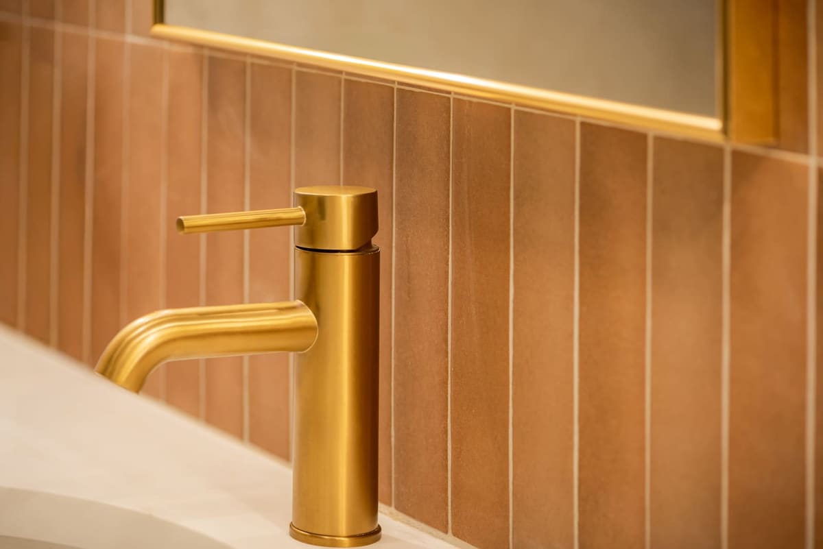 Brushed brass faucet against vertical terracotta tile below an arched mirror