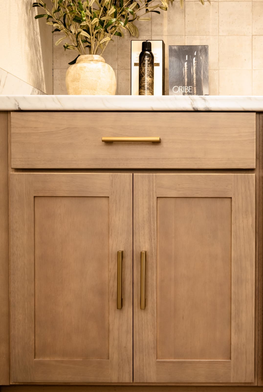 Light oak shaker cabinet with brass pulls below a tiled wall and a vase of olive branches
