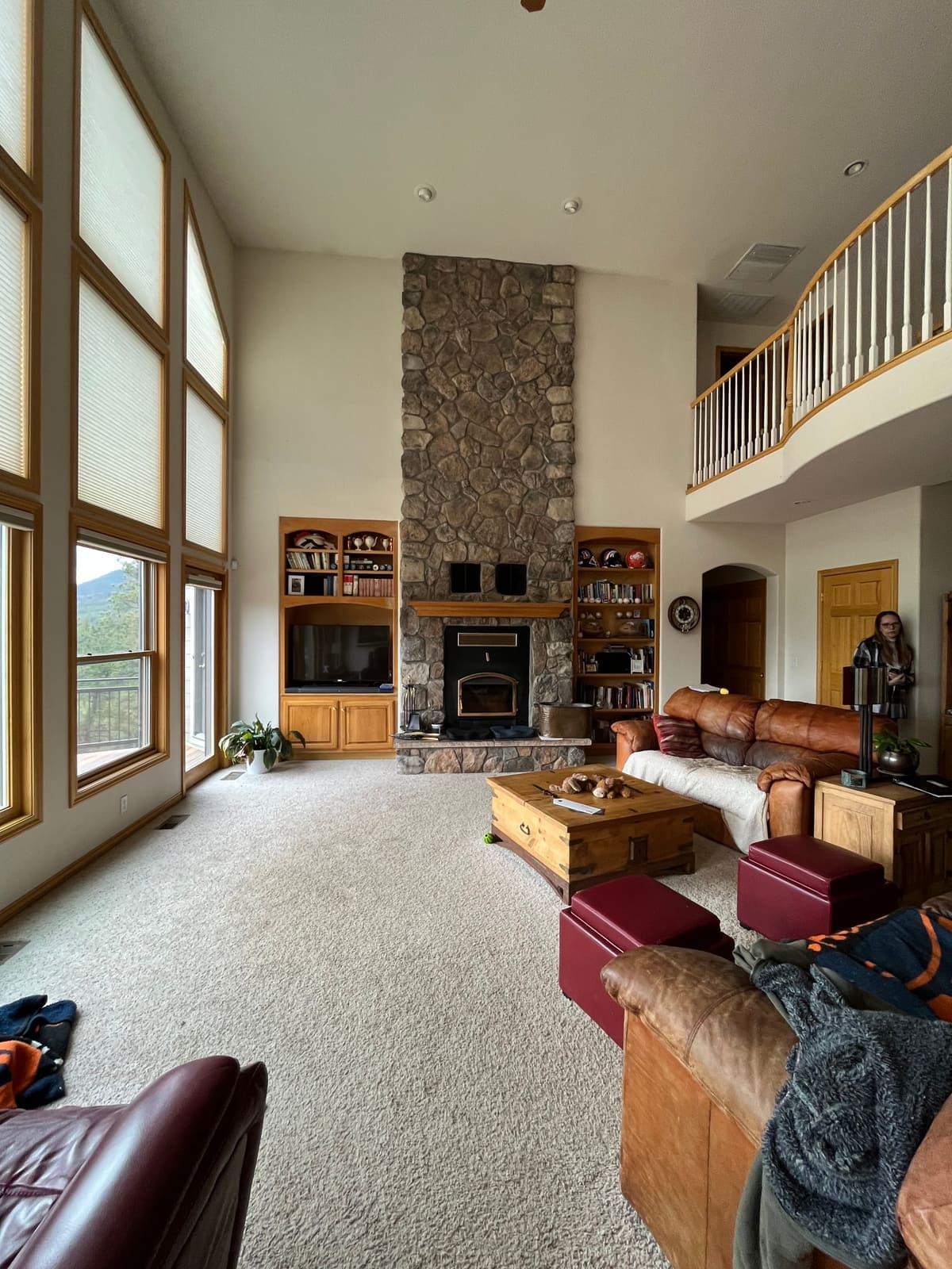 Before the remodel: tall living room with a loft balcony railing above dated oak trim and stone fireplace