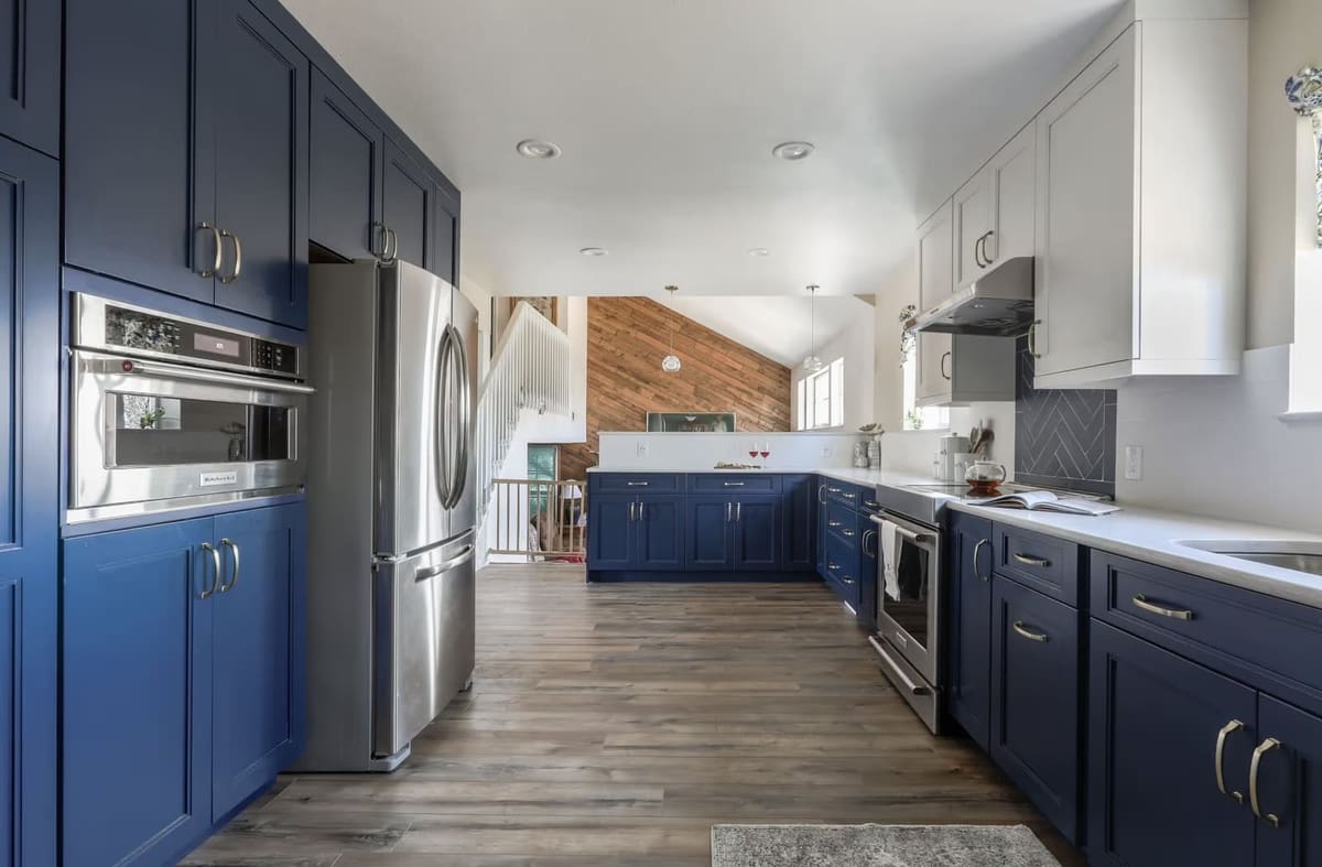 Galley view of a kitchen with a navy cabinet wall around the fridge and wall oven, white uppers and wood-look floors