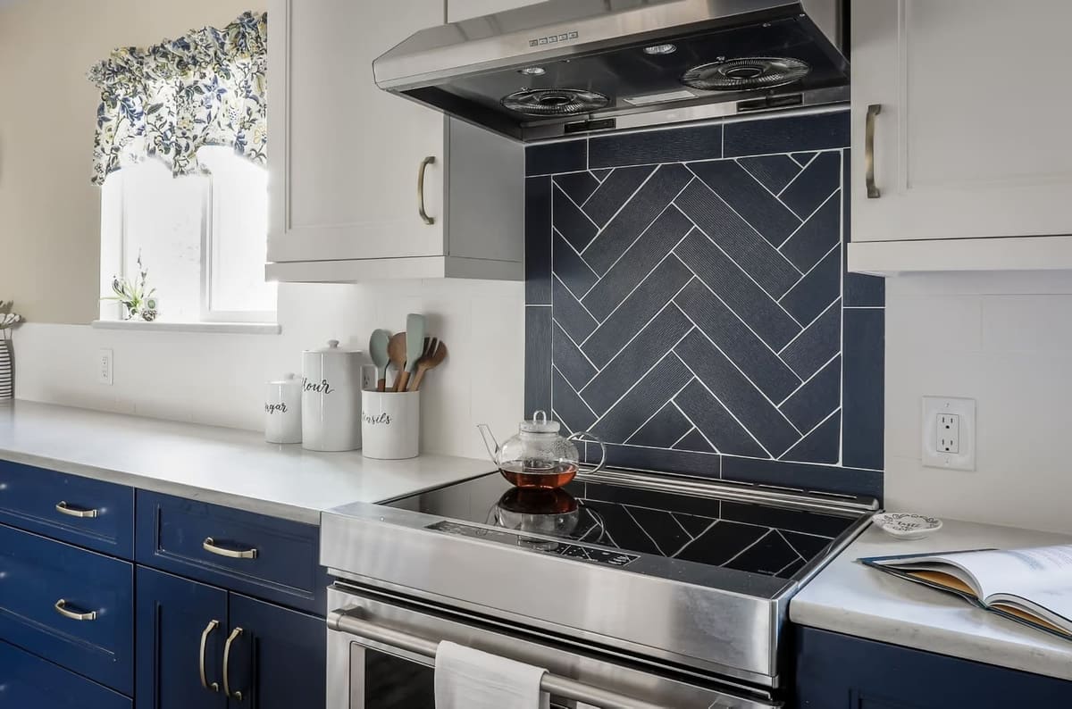 Range wall with a navy herringbone tile panel, stainless hood, white upper cabinets and navy drawers with brass pulls