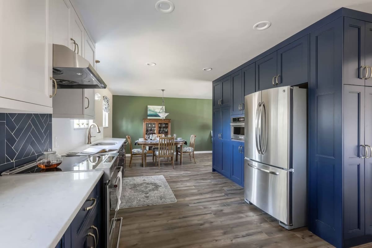 Kitchen with a tall navy pantry wall and stainless fridge opening into a dining room with a green wall and oak table