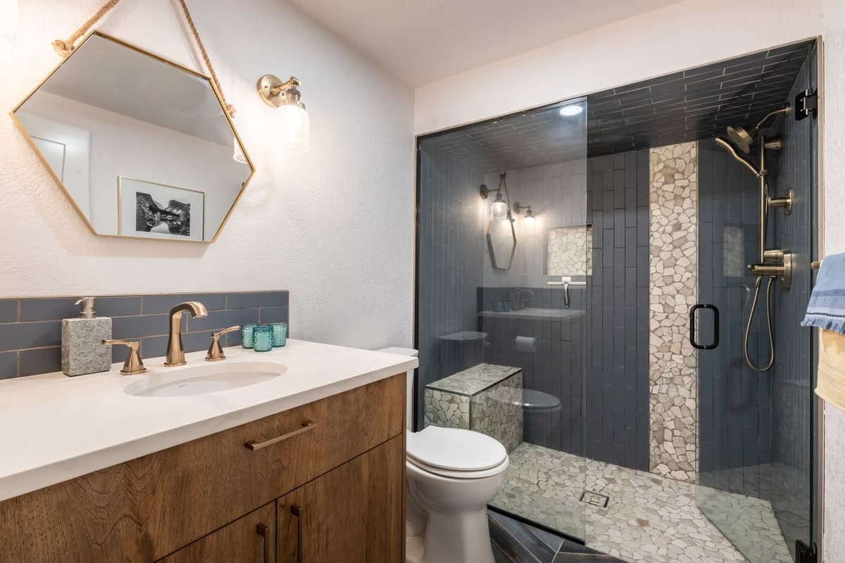Bathroom with walnut vanity, brass fixtures, and walk-in shower in slate blue tile with pebble mosaic stripe