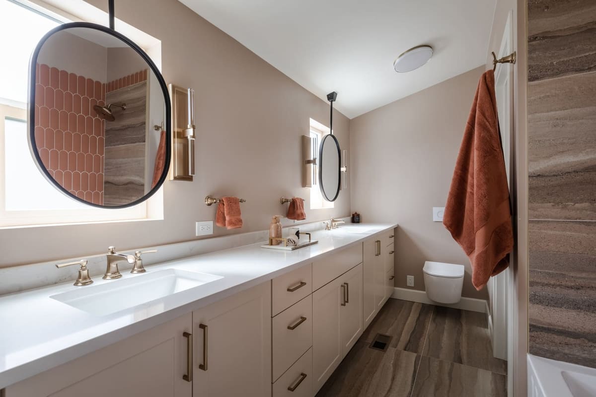 Long cream shaker vanity with champagne bronze faucets, oval hanging mirrors and a wall-hung toilet