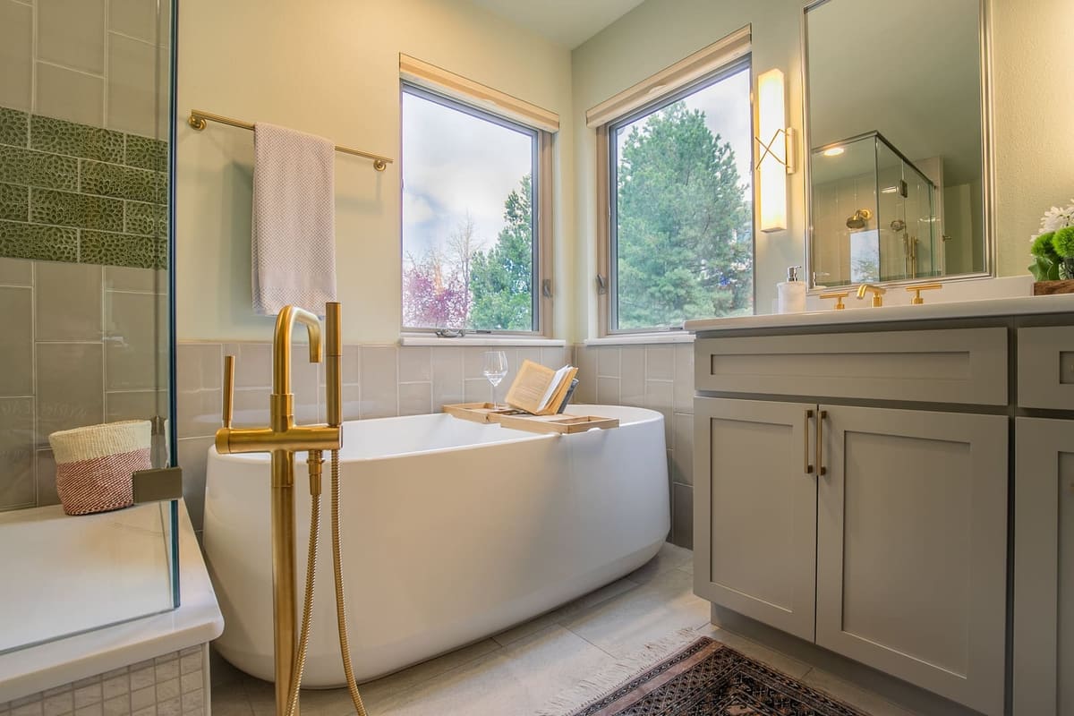 Primary bath corner with a freestanding tub between two windows, brass tub filler and gray shaker vanity