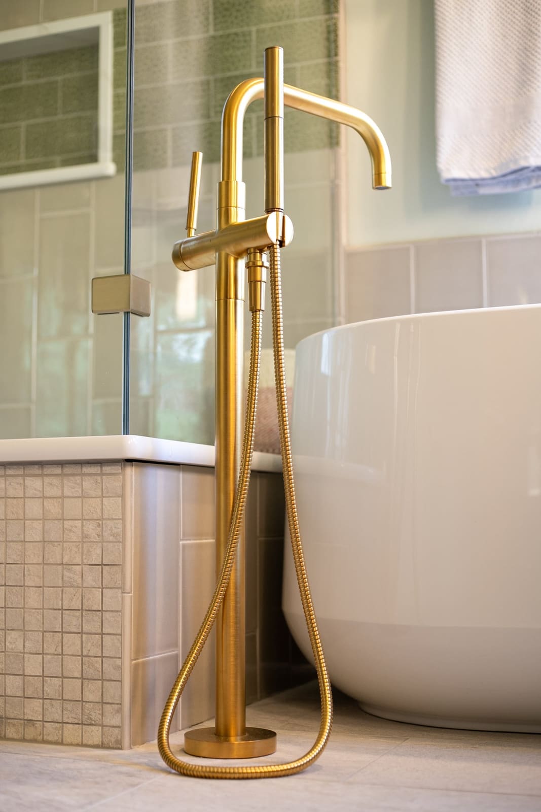 Brushed brass floor-mounted tub filler beside a freestanding tub, with sage leaf tile in the shower behind