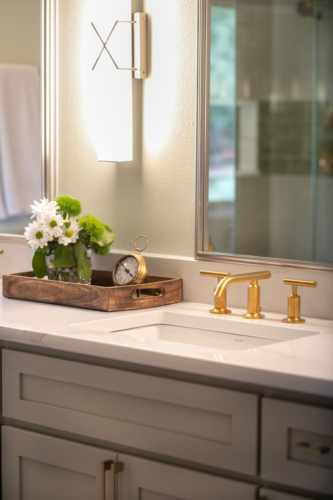 Vanity detail with a brushed brass faucet, white quartz top, gray shaker drawers and an X-detail sconce