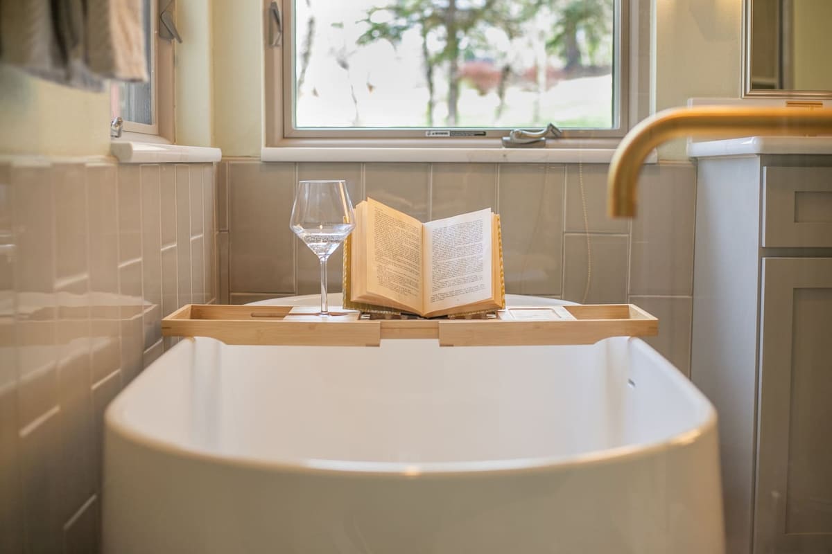 Bamboo tub tray with a wine glass and open book across a freestanding tub below a garden window