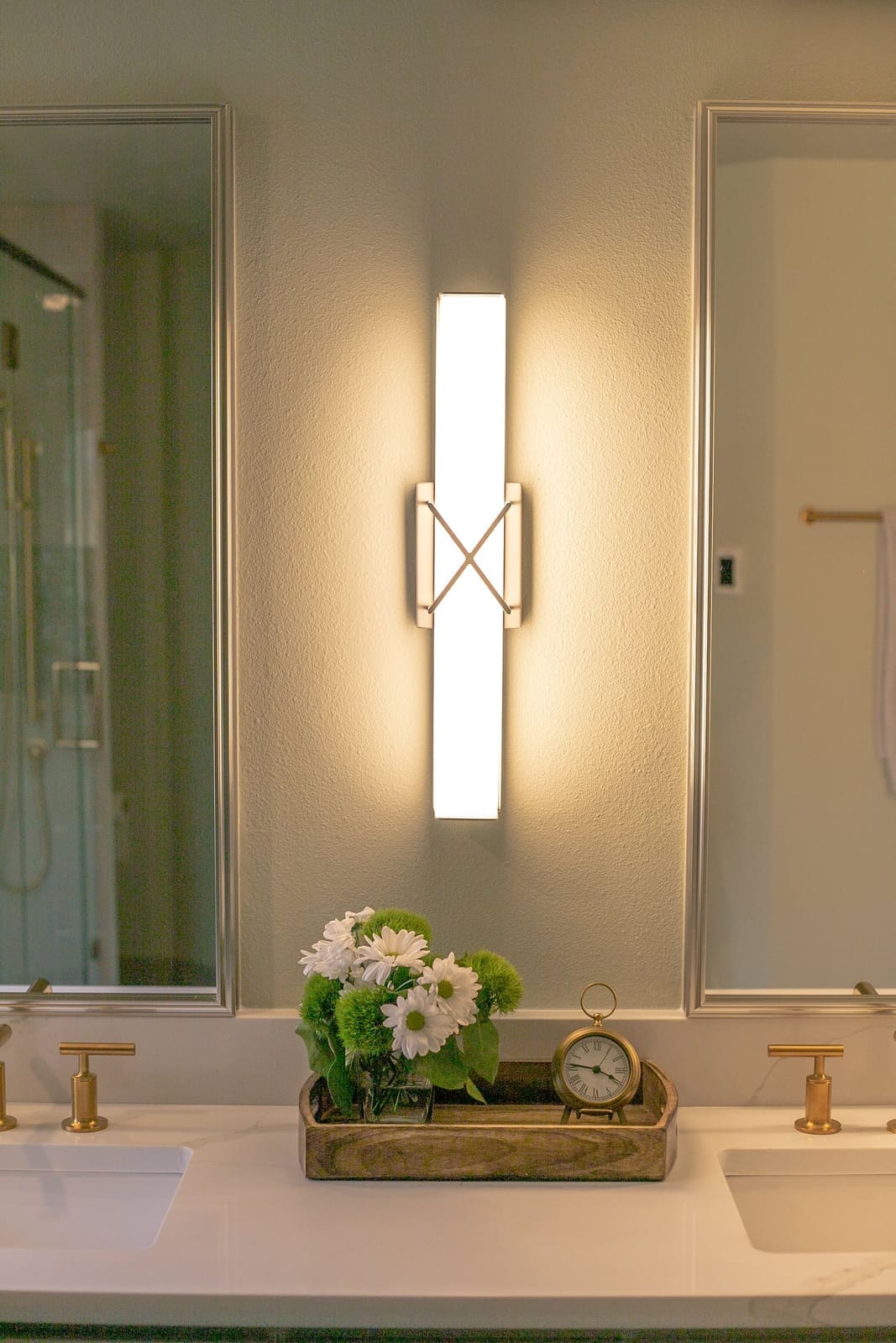 Glowing X-detail wall sconce between two framed mirrors above a tray of daisies and a brass clock