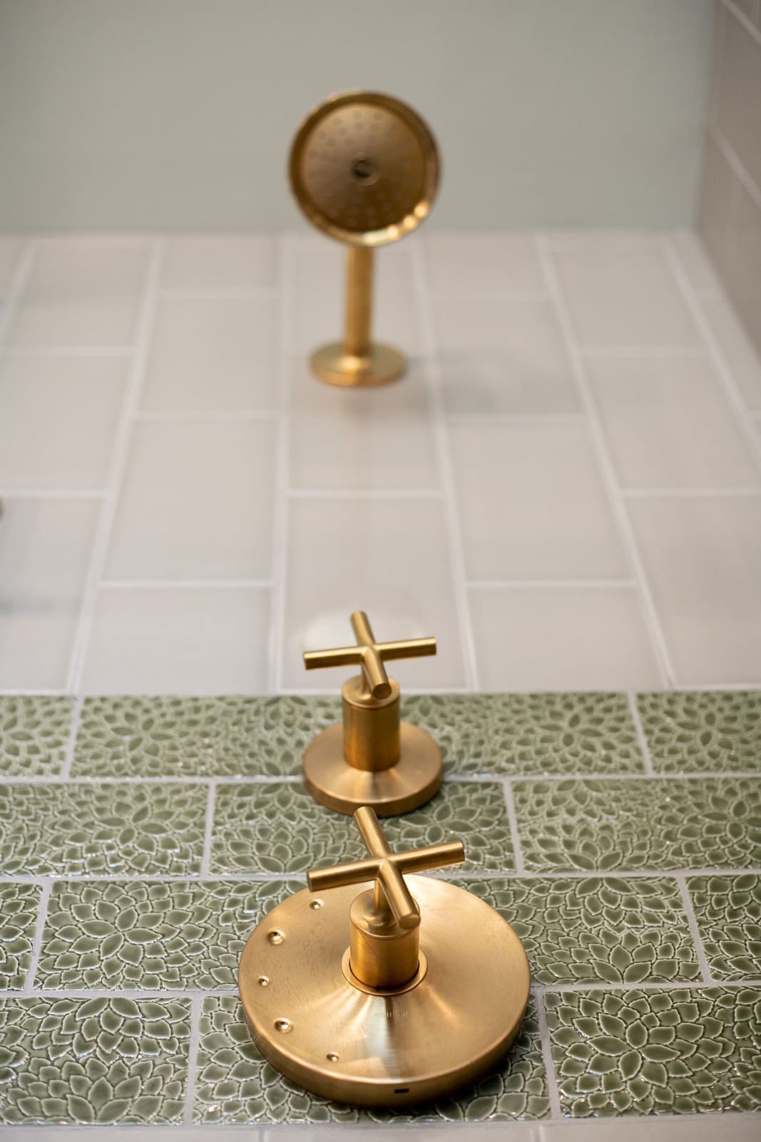 Brushed brass cross-handle tub valve and handshower on sage green leaf-texture tile and white tile