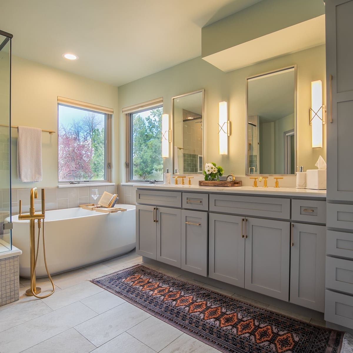 Primary bath with a glass shower, freestanding tub, brass floor filler, gray double vanity and patterned runner