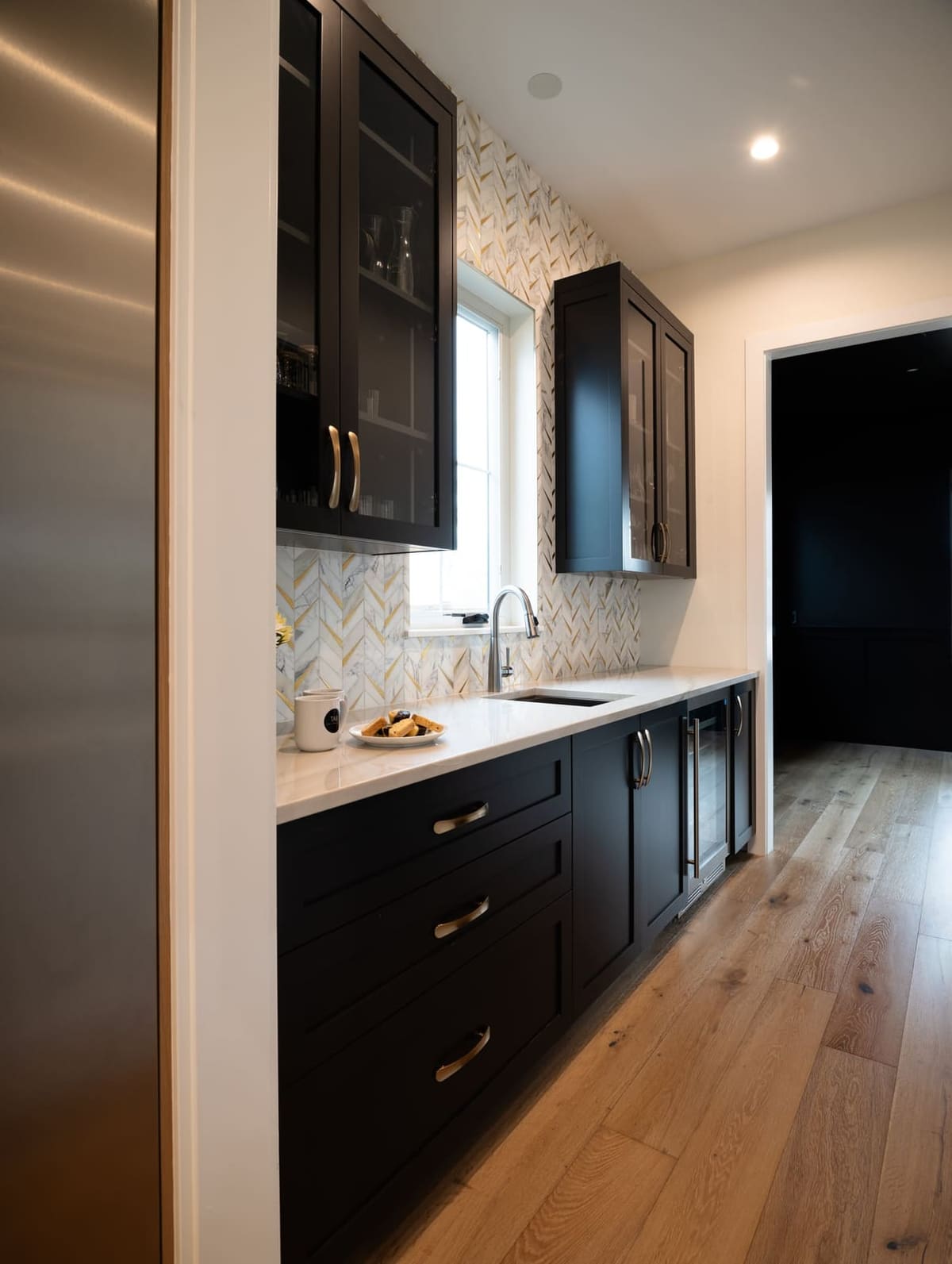 Butler's pantry with black shaker cabinets, glass uppers, marble and brass chevron tile and wide oak floors