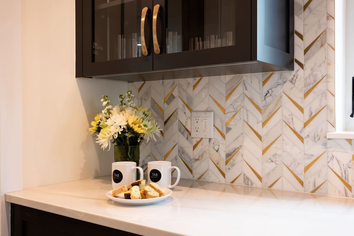 Black glass-front cabinet with champagne arched pulls over a marble and brass chevron backsplash