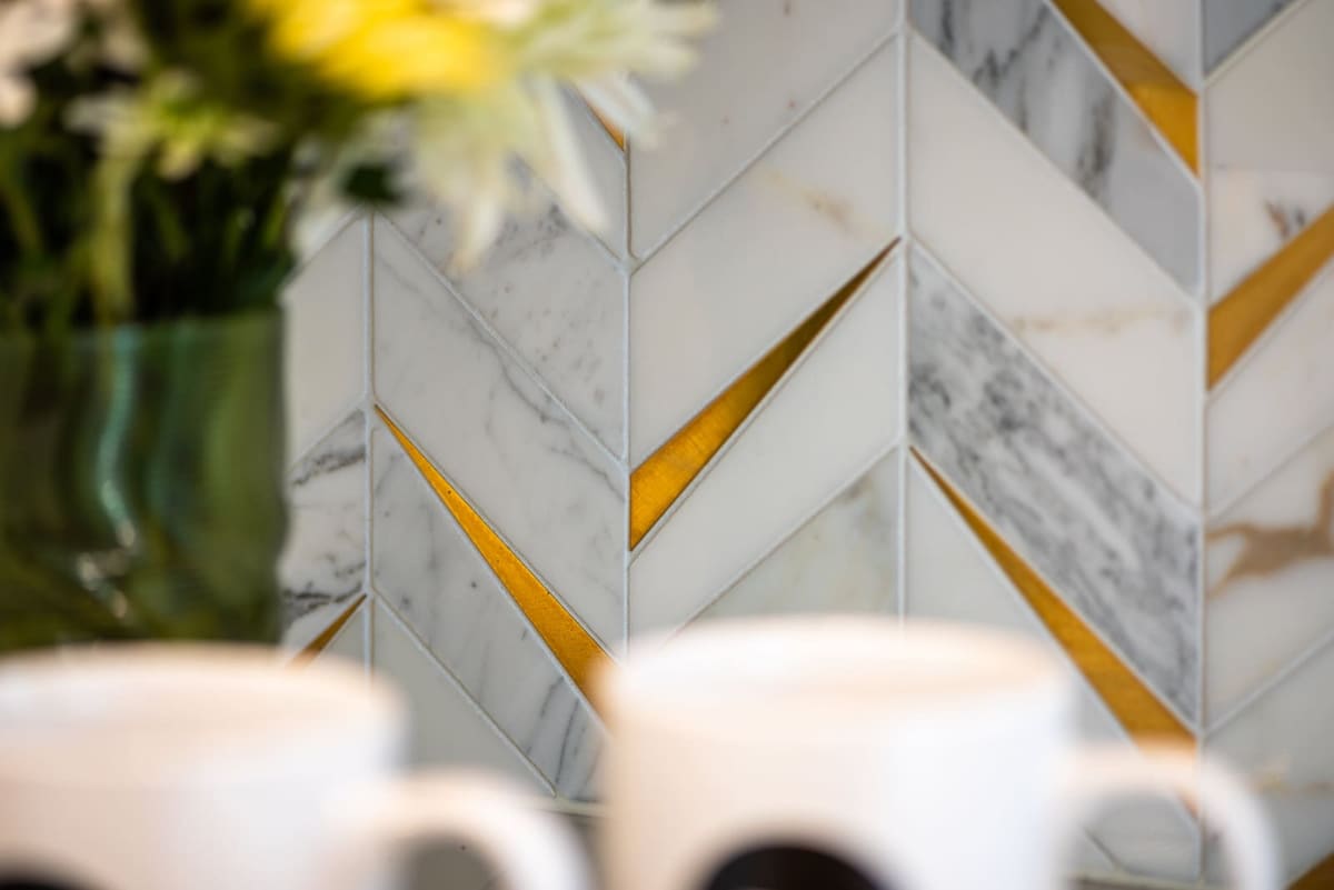 Macro view of white marble chevron tile with polished brass inlay strips, flowers and mugs blurred