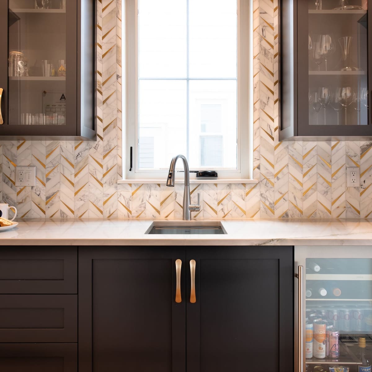 Butler's pantry centered on a window, with marble and brass chevron tile, glass uppers and a beverage fridge