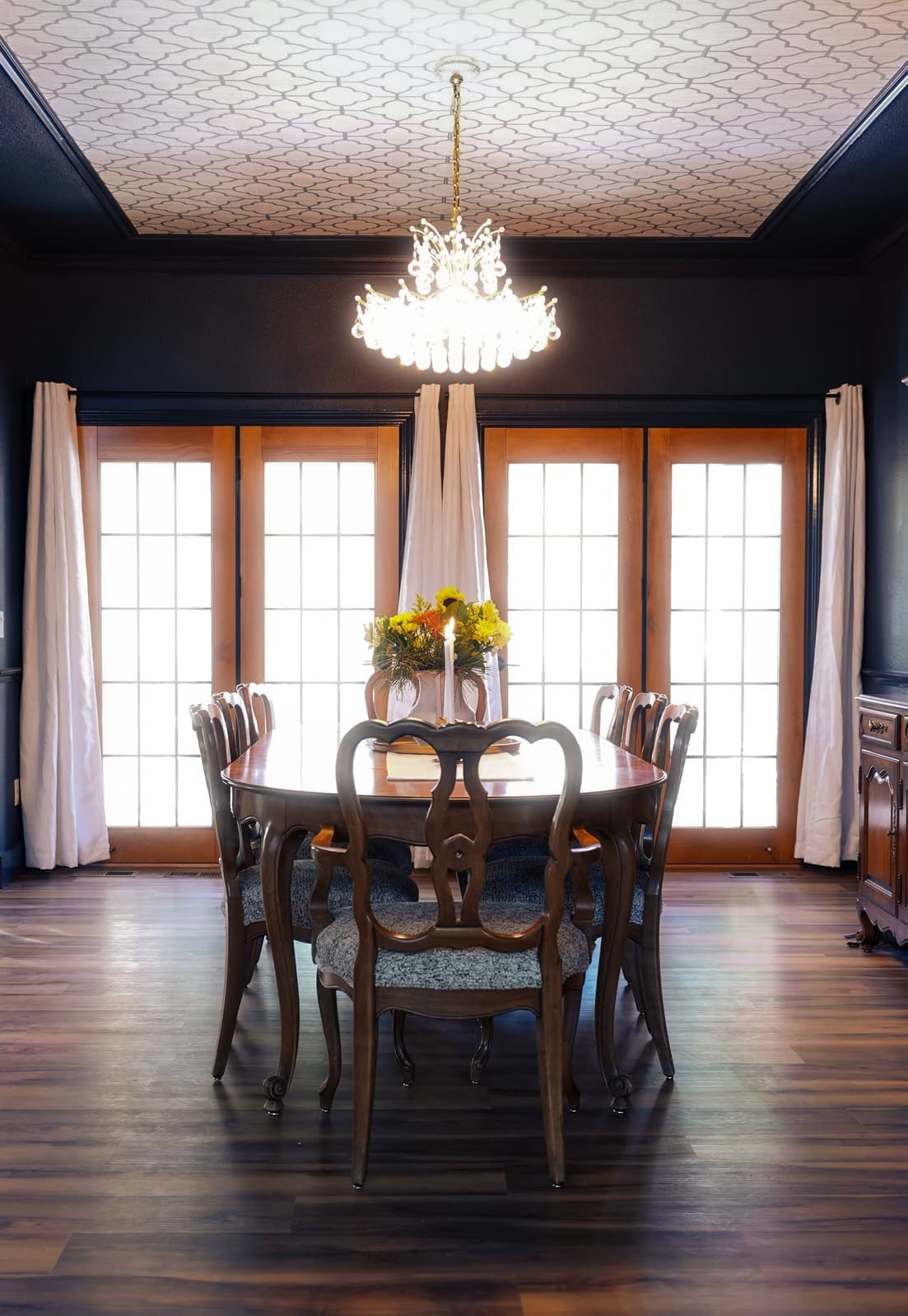Dining room with a wallpapered ceiling, crystal chandelier and French doors dressed in white drapes