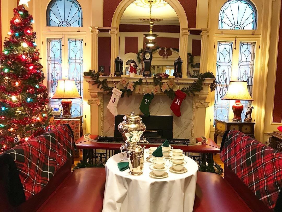 Tree with red and gold ornaments beside a mantel hung with stockings, plaid-covered seating and a silver urn