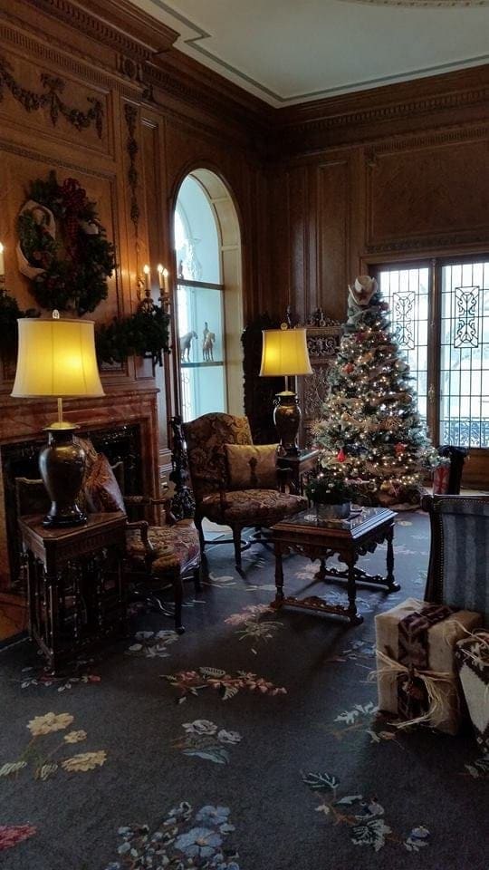 Wood-paneled library with a lit Christmas tree, a wreath over the fireplace and glowing table lamps
