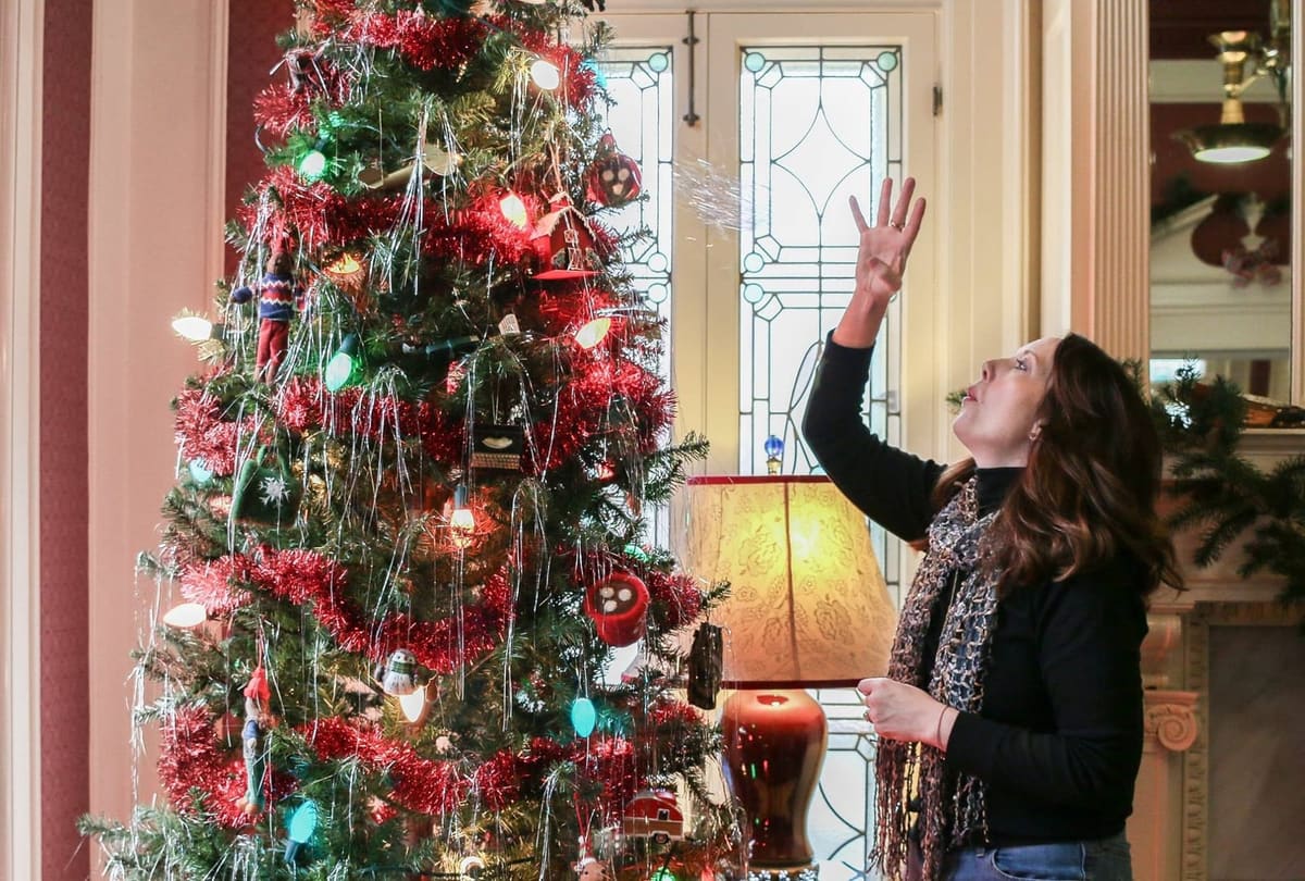 A TAB designer hanging ornaments on a tinsel-wrapped Christmas tree beside leaded glass doors