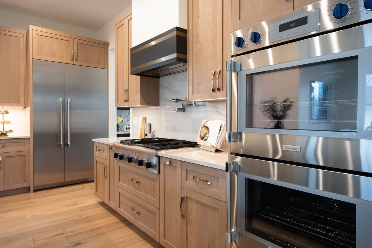 White oak cabinetry around a stainless double wall oven, range top and built-in refrigerator