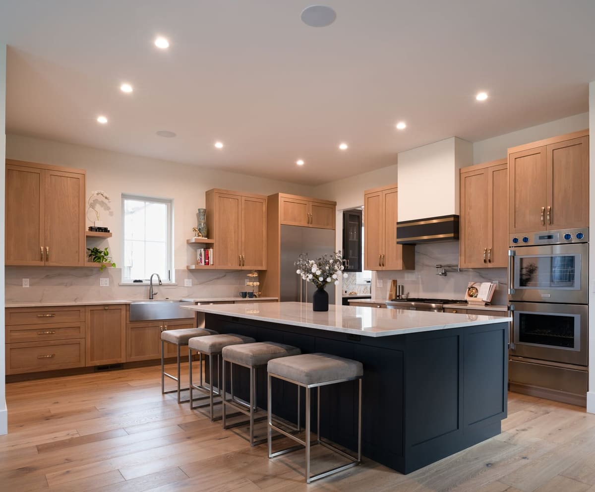 Navy island and white oak cabinets under recessed lights with a farmhouse sink below the window