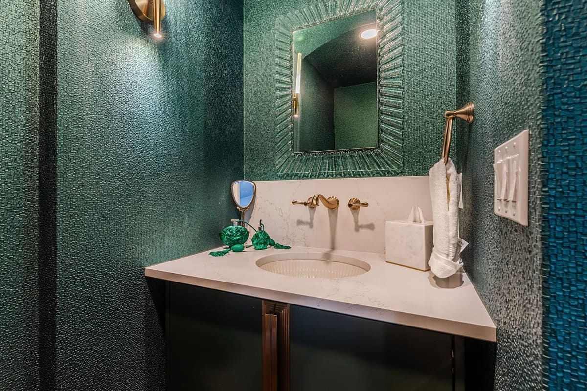 Powder room with emerald textured wallcovering, a sunburst mirror, white stone vanity and brass faucet