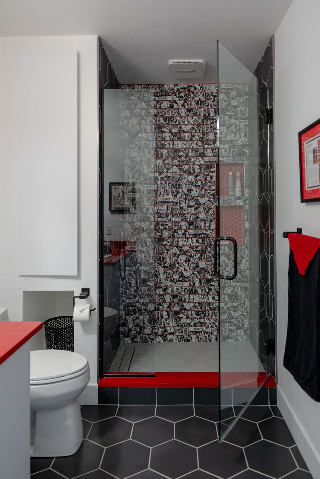 Walk-in shower with graffiti-print subway tile, black hexagon floor tile, a red curb and frameless glass
