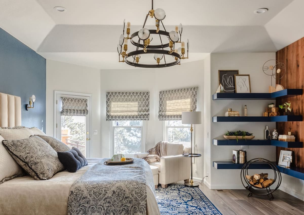 Primary bedroom with a wagon-wheel chandelier over the bed and a bay window with Roman shades