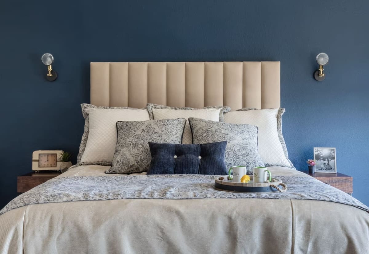 Cream channel headboard and patterned bedding against a navy wall between brass sconces