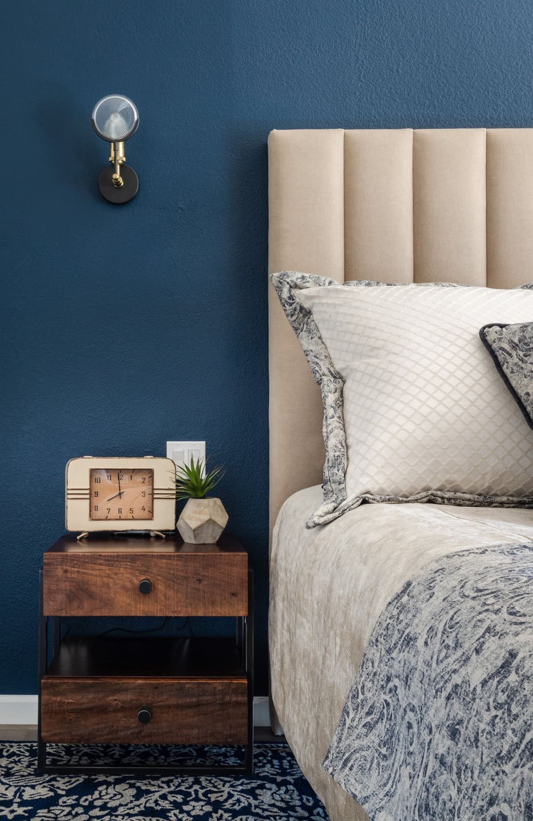 Bedside vignette with a cream channel headboard, navy wall, brass sconce and wood nightstand