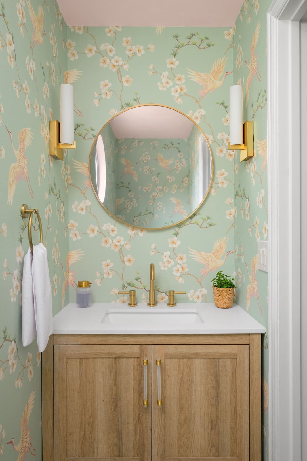 Powder room with mint crane and blossom wallpaper, a round brass mirror, brass sconces and light oak vanity