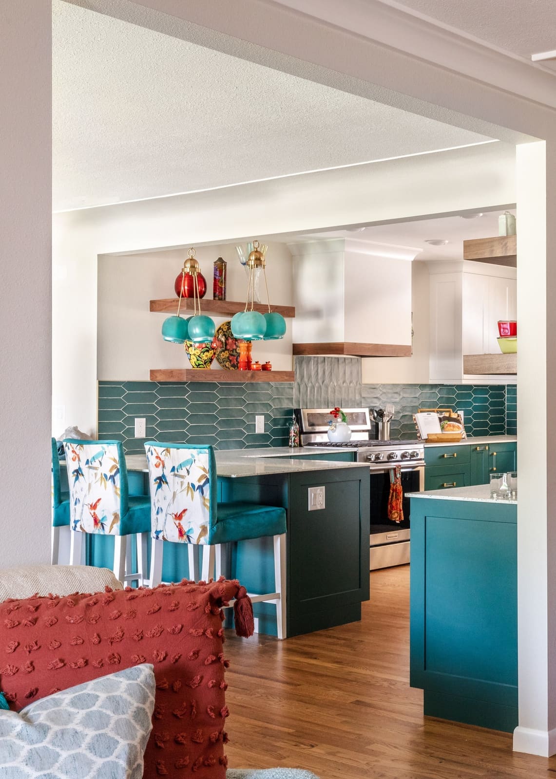 View into the teal kitchen with floral bar stools at the island and walnut floating shelves