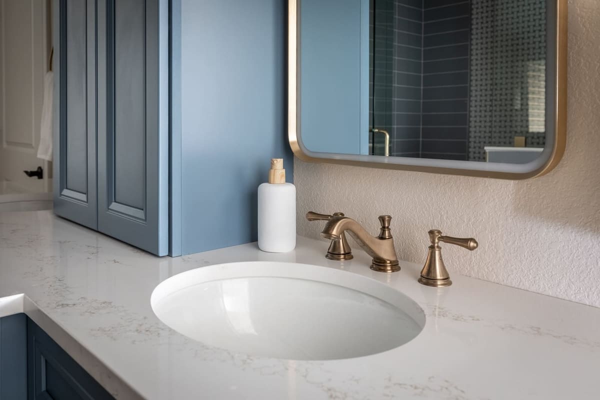 Brass faucet on white quartz beside a blue linen tower and brass-framed mirror