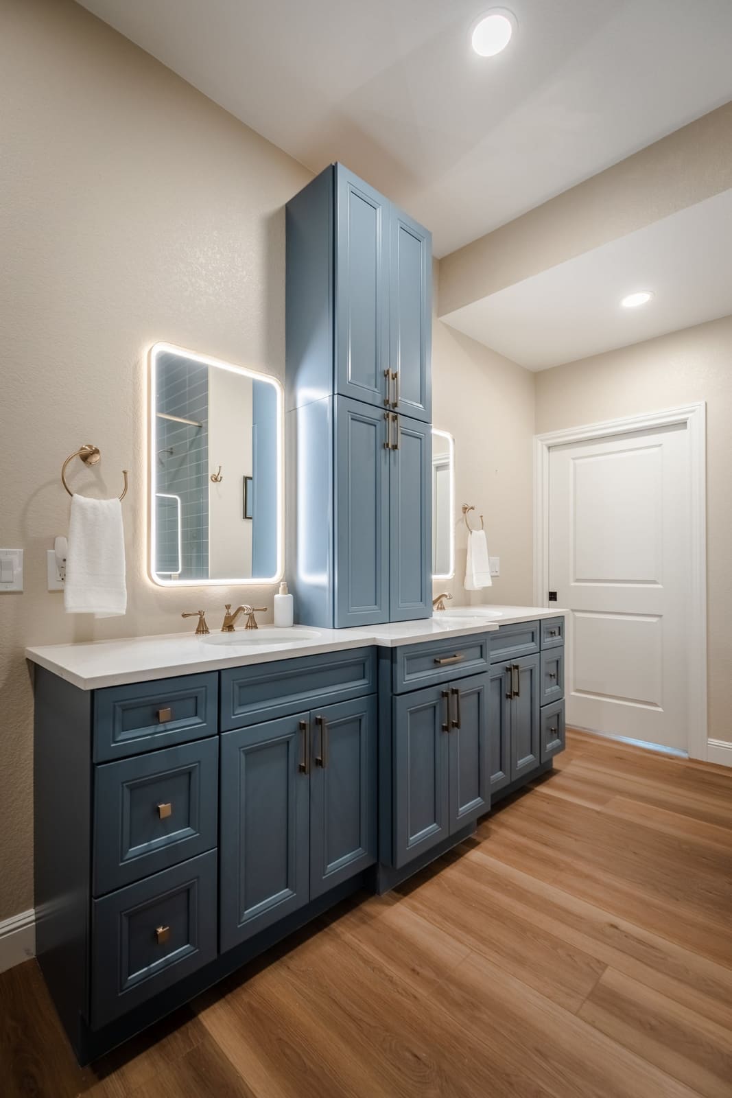 Blue double vanity and linen tower with backlit mirrors beside a white door