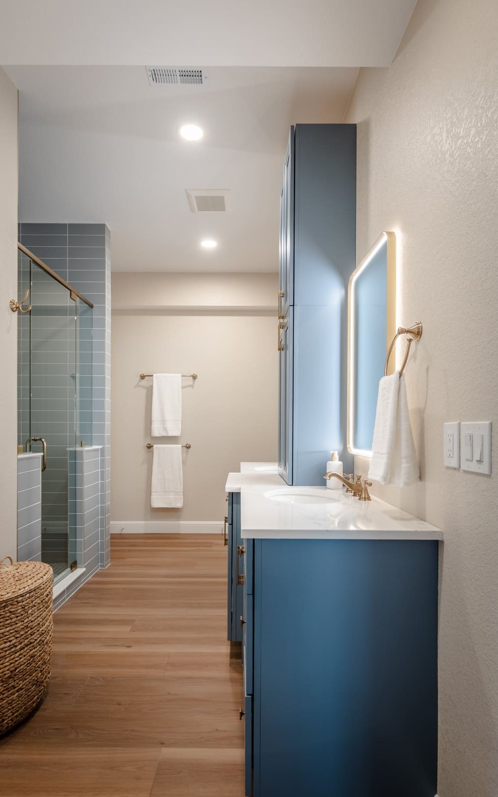 View down the bath past the blue vanity and backlit mirror toward the shower