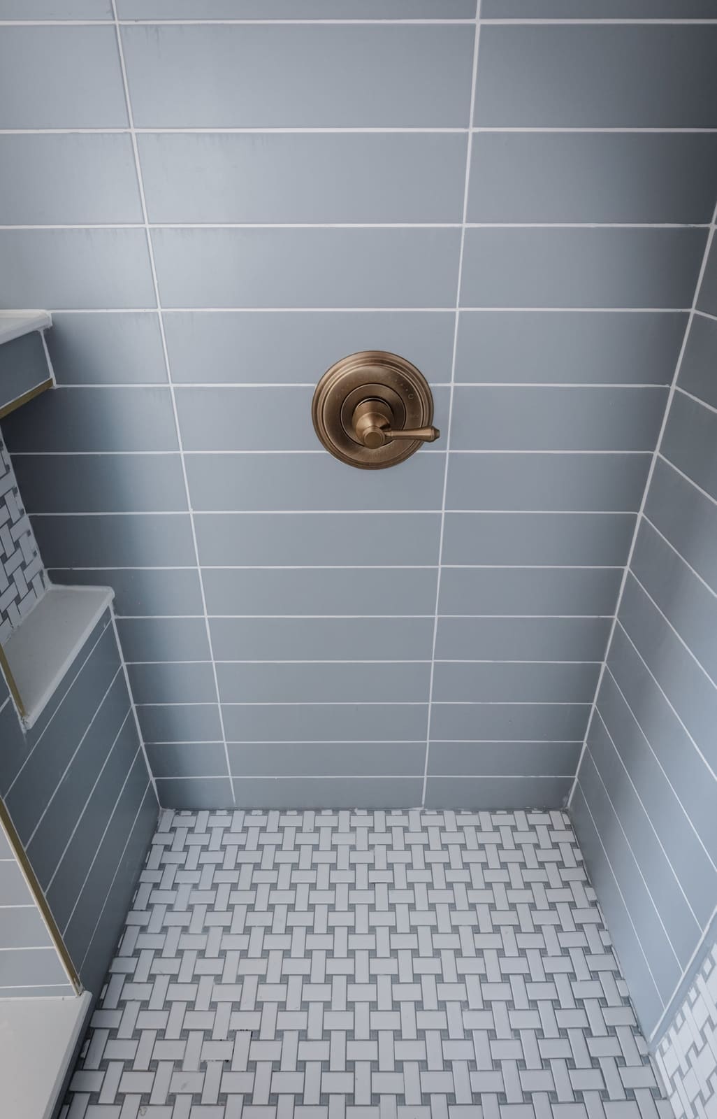 Shower wall in gray-blue tile with a brass valve above a white mosaic floor