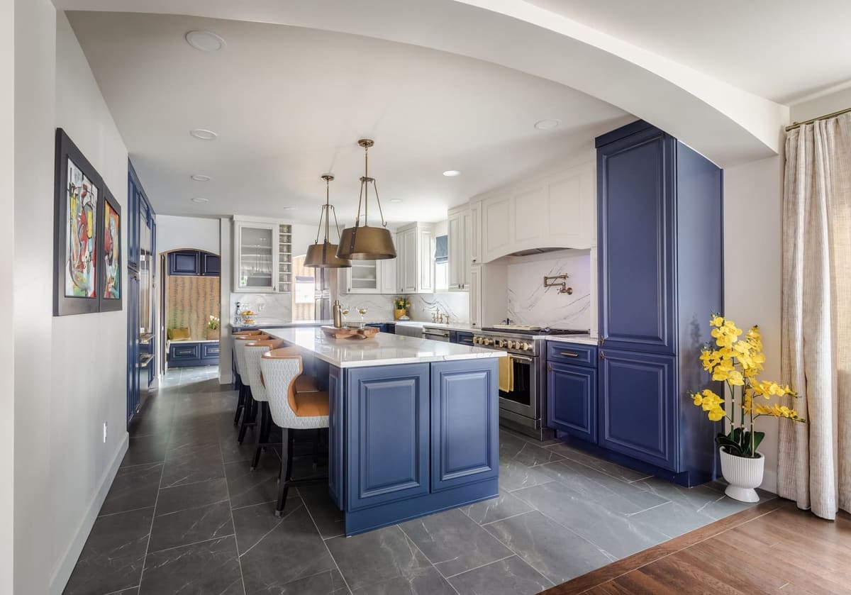 Kitchen with a navy raised-panel island, white uppers and brass pendants, seen through an arched opening