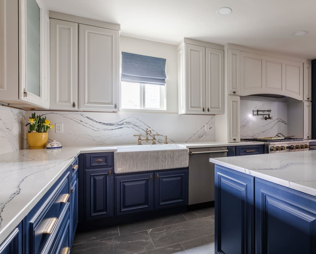 White uppers and navy lowers around a farmhouse sink with a marble-look quartz backsplash