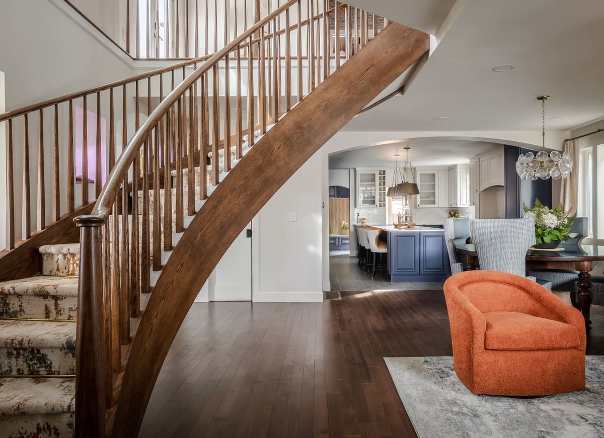 Curved wood staircase leading to an orange swivel chair, the kitchen beyond
