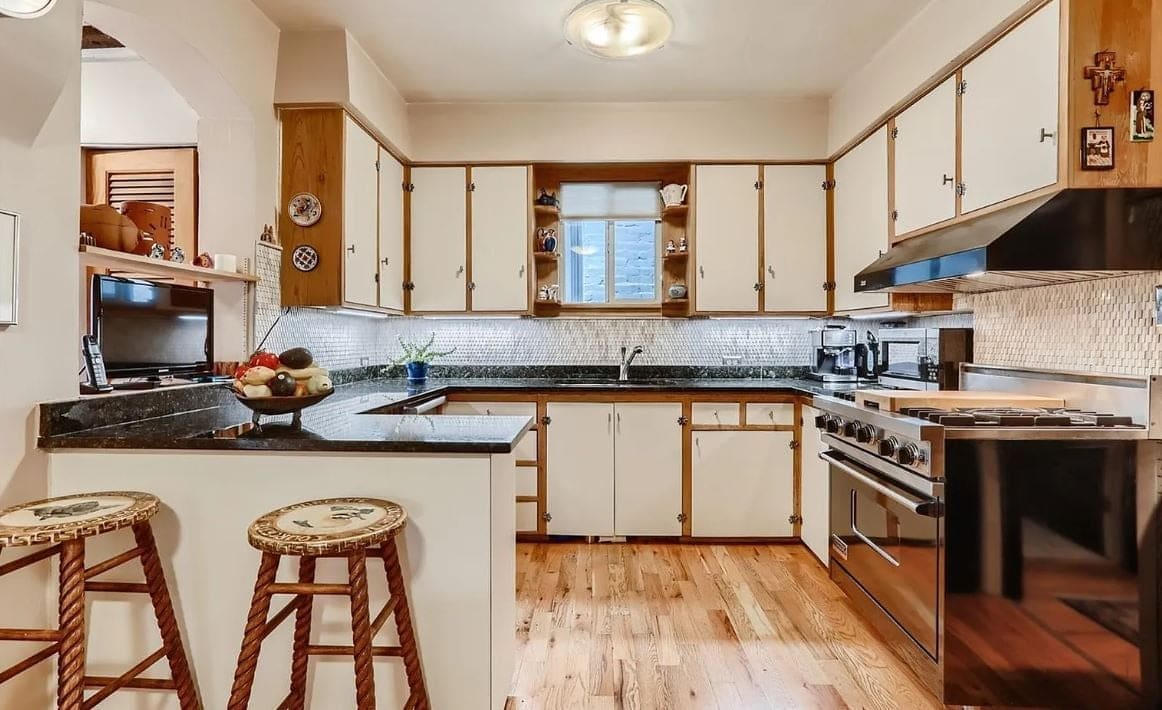 Before the remodel: cream and wood-trimmed kitchen with black granite, a pro range and wicker-seat stools