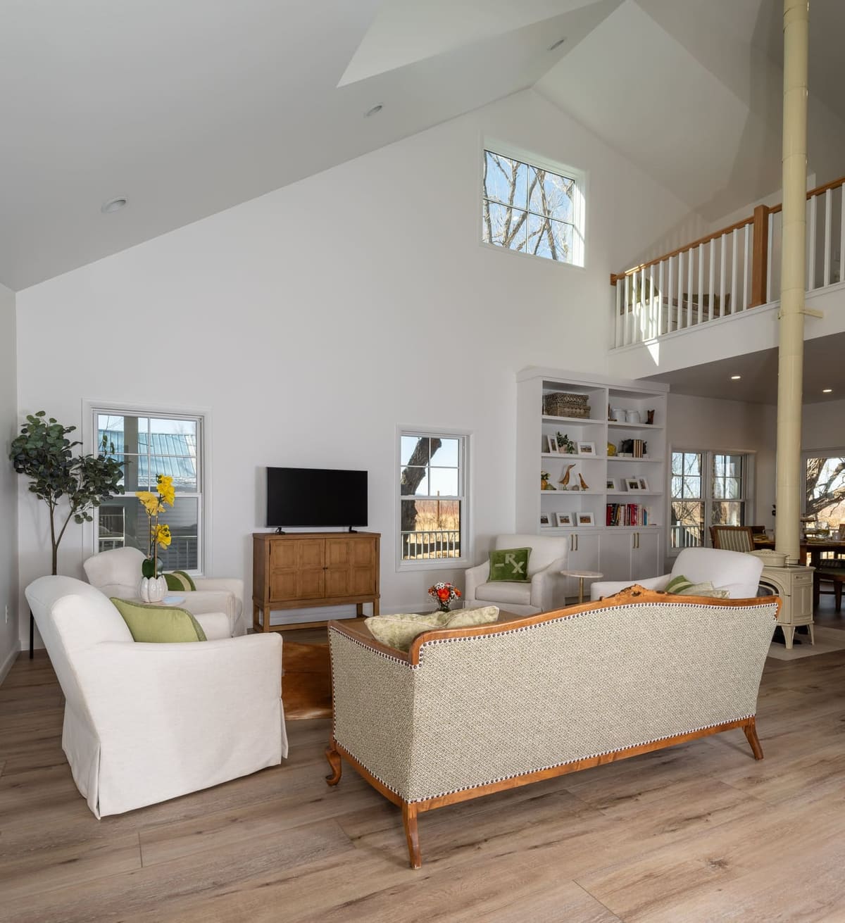 Vaulted living room with white slipcovered chairs, a caned sofa and a loft railing above