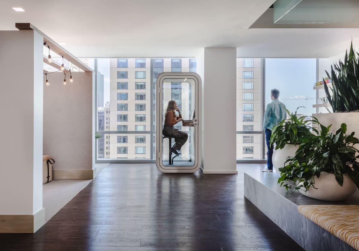 Bright office corridor with a phone booth pod, large planters and tall windows onto the city