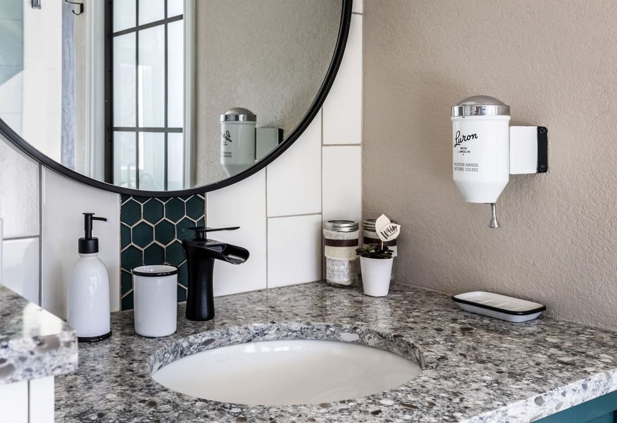 Vanity detail with a round black mirror, granite counter, matte black faucet and green hex tile