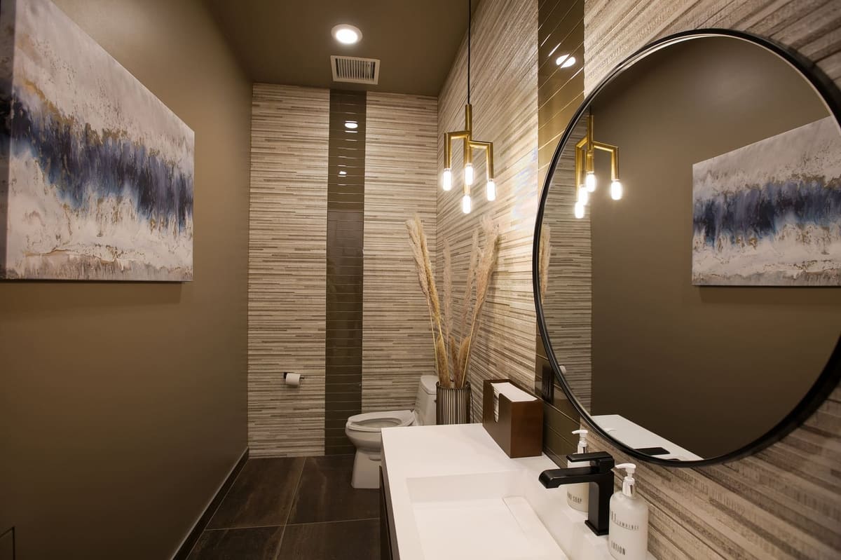 Restroom with dark walls, a band of sand-toned stone tile, round black mirror and white trough sink