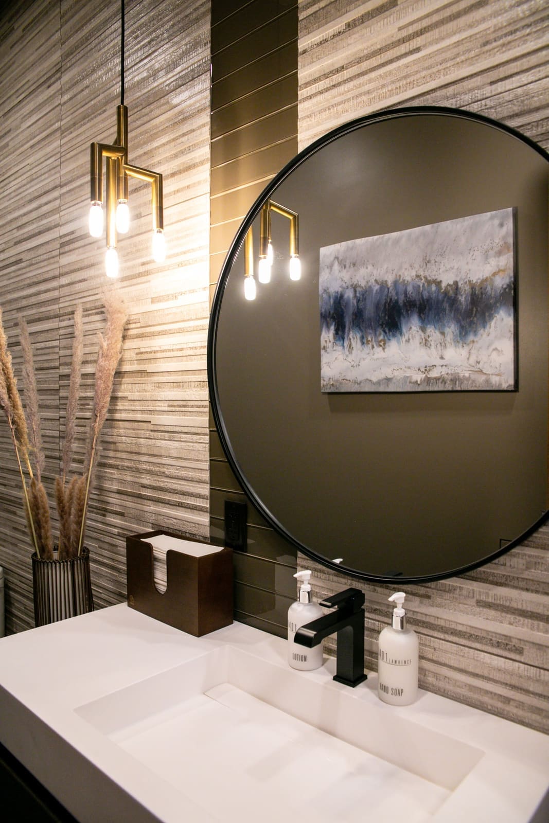 Restroom vanity with a round black mirror, gold cluster pendant and sand-toned stone tile
