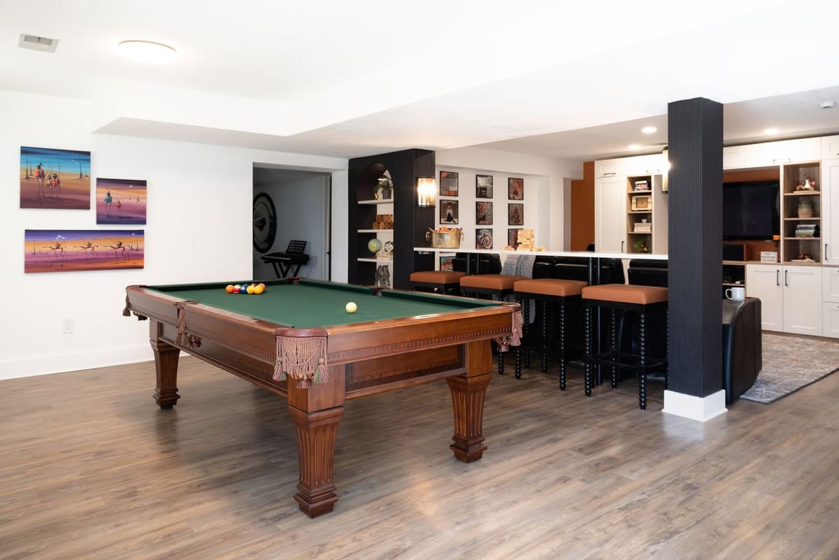 Finished basement game area with a green felt pool table, colorful art, a fluted black column and bar seating