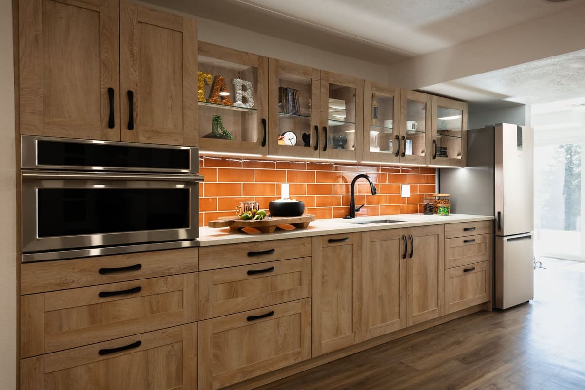 Basement kitchenette in light oak shaker cabinetry with a wall oven, orange glazed tile and quartz counters