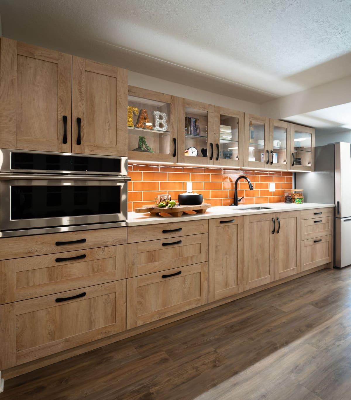 Angled view of a basement kitchenette with oak shaker cabinets, glass uppers and an orange subway backsplash