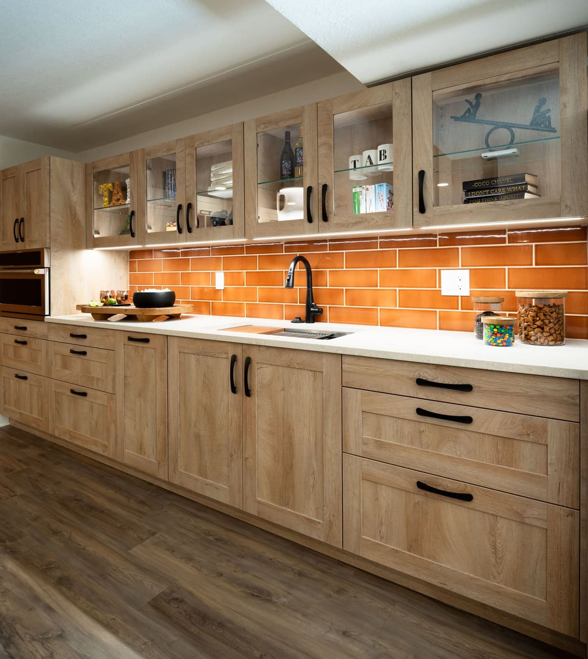 Angled kitchenette view with glass-front oak uppers holding mugs and books over orange subway tile