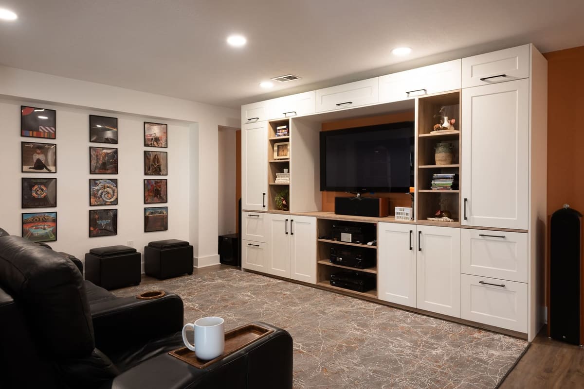 Basement media wall of white shaker cabinets and oak open shelves around a TV with an orange back panel
